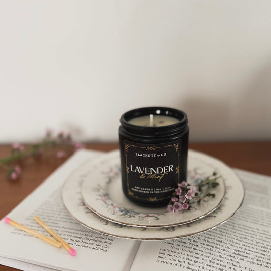Lavender & Mint scented natural soy candle in a black candle jar by Blackett & Co. on a decorative plate with flowers and an open book