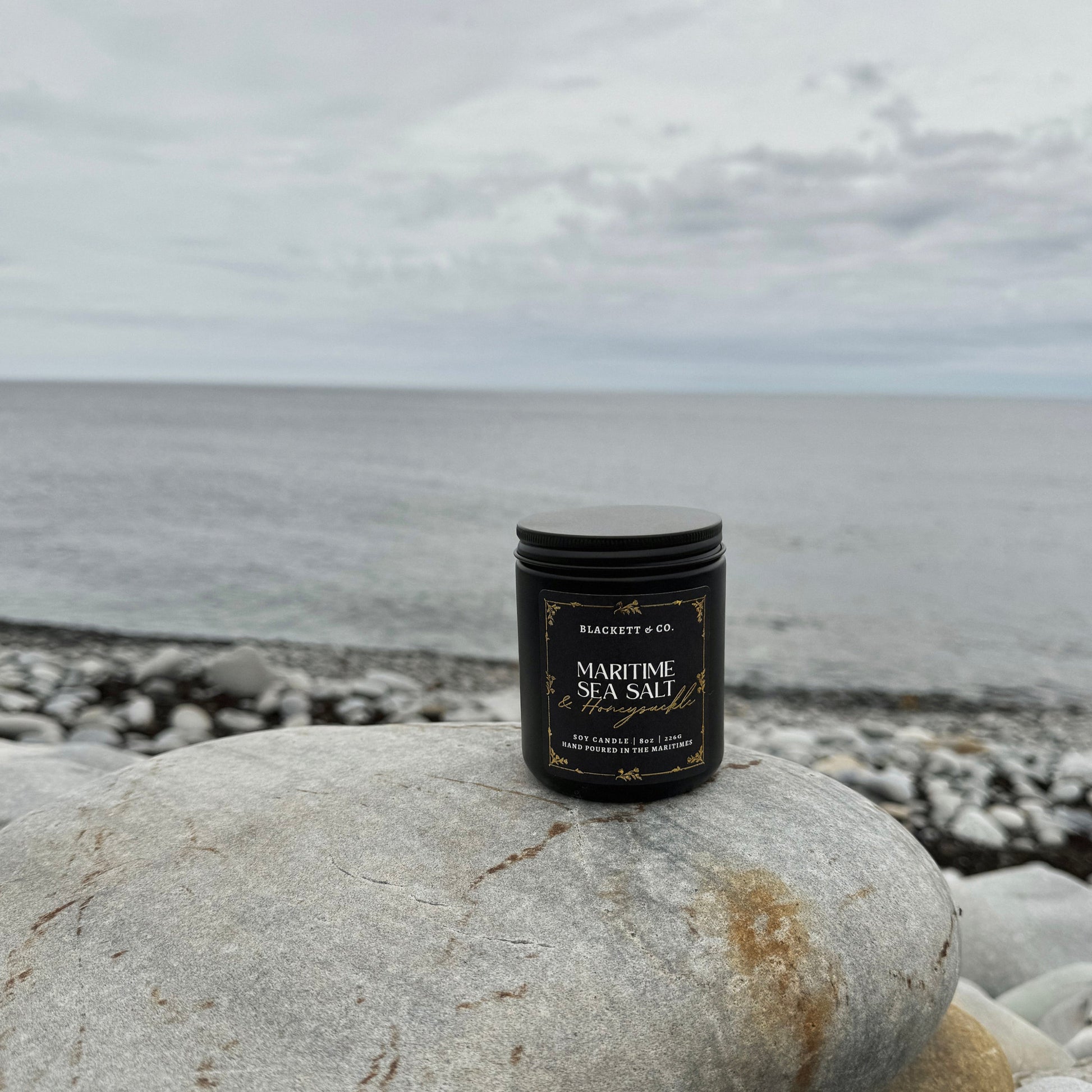 Maritime Sea Salt & Honeysuckle scented natural soy candle on a pebble beach with ocean and sky in the background