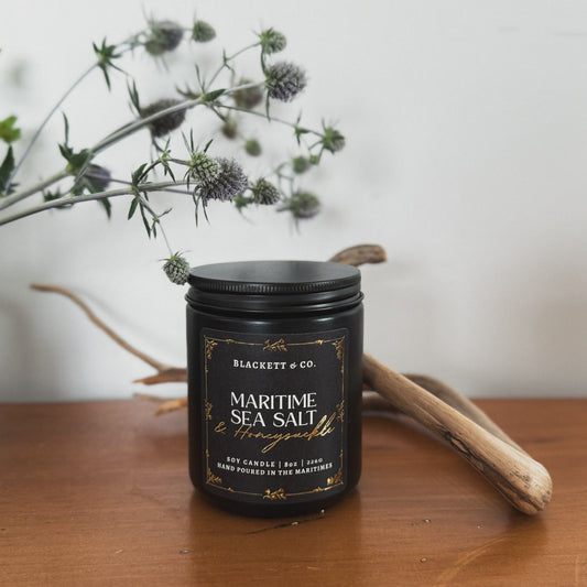 Maritime Sea Salt & Honeysuckle scented natural soy candle in a black jar by Blackett & Co. on a wooden surface with thistle and driftwood in the background