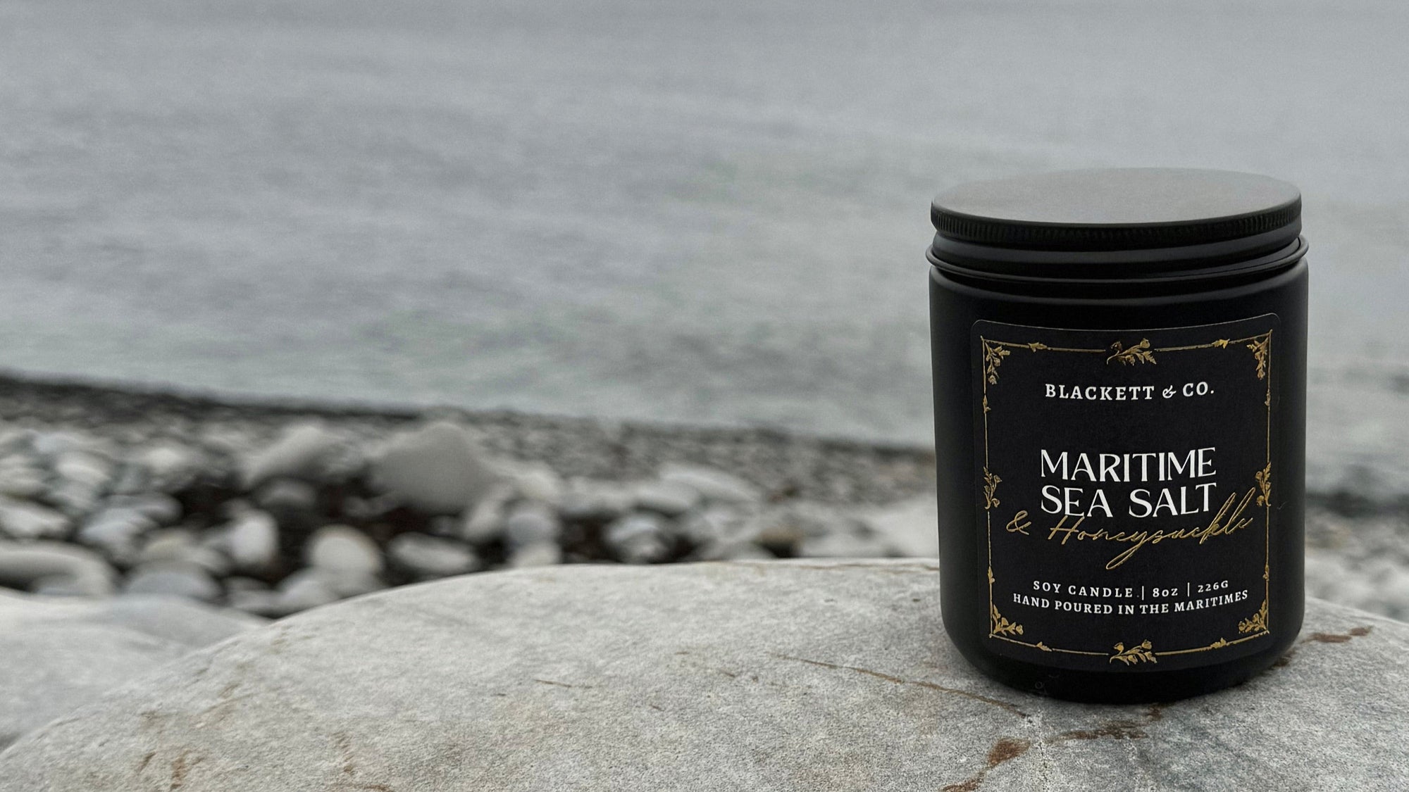 Maritime Sea Salt & Honeysuckle scented natural soy candle in a black jar by Blackett & Co. on a rocky beach.