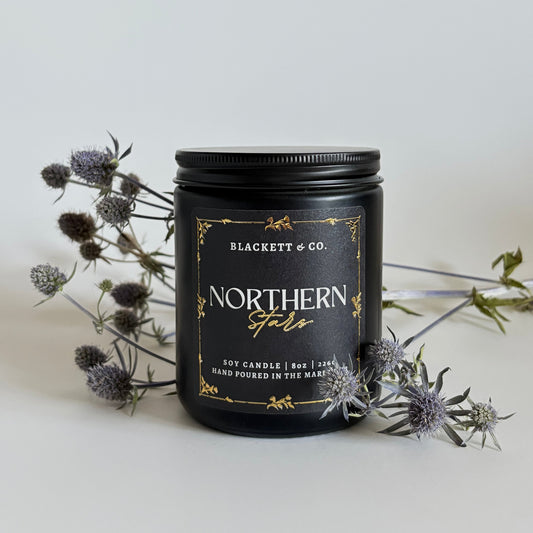 Northern Stars scented natural soy candle in a black jar by Blackett & Co. with a light background and thistles