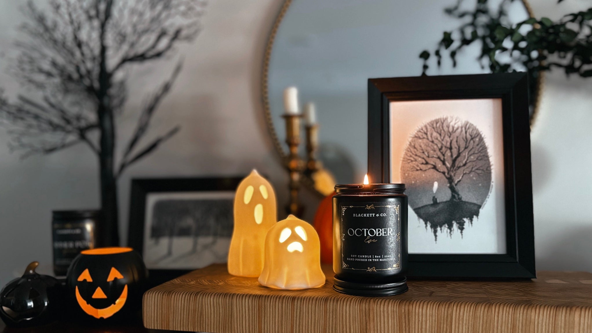 Halloween-themed decor with an October Eve natural scented soy candle by Blackett & Co. and ghost-shaped ceramic lights on a wooden surface.