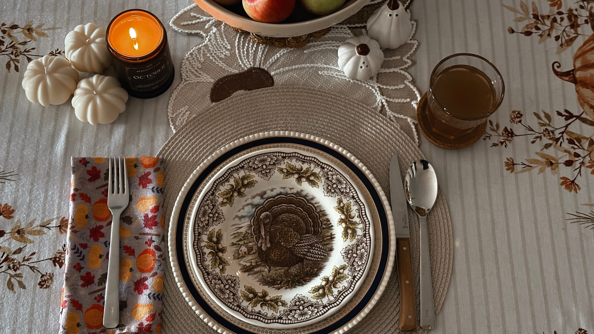 Thanksgiving table setting with a bowl of apples, October Eve scented soy candle, and decorative items on a patterned tablecloth.