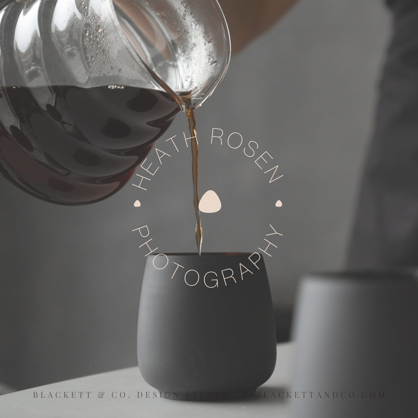 Photography brand logo semi-transparent circular watermark centred over an image of a dark grey ceramic mug while coffee is being poured from a clear glass carafe.