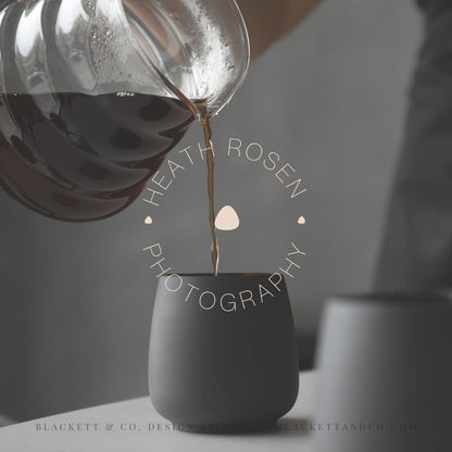 Photography brand logo semi-transparent circular watermark centred over an image of a dark grey ceramic mug while coffee is being poured from a clear glass carafe.
