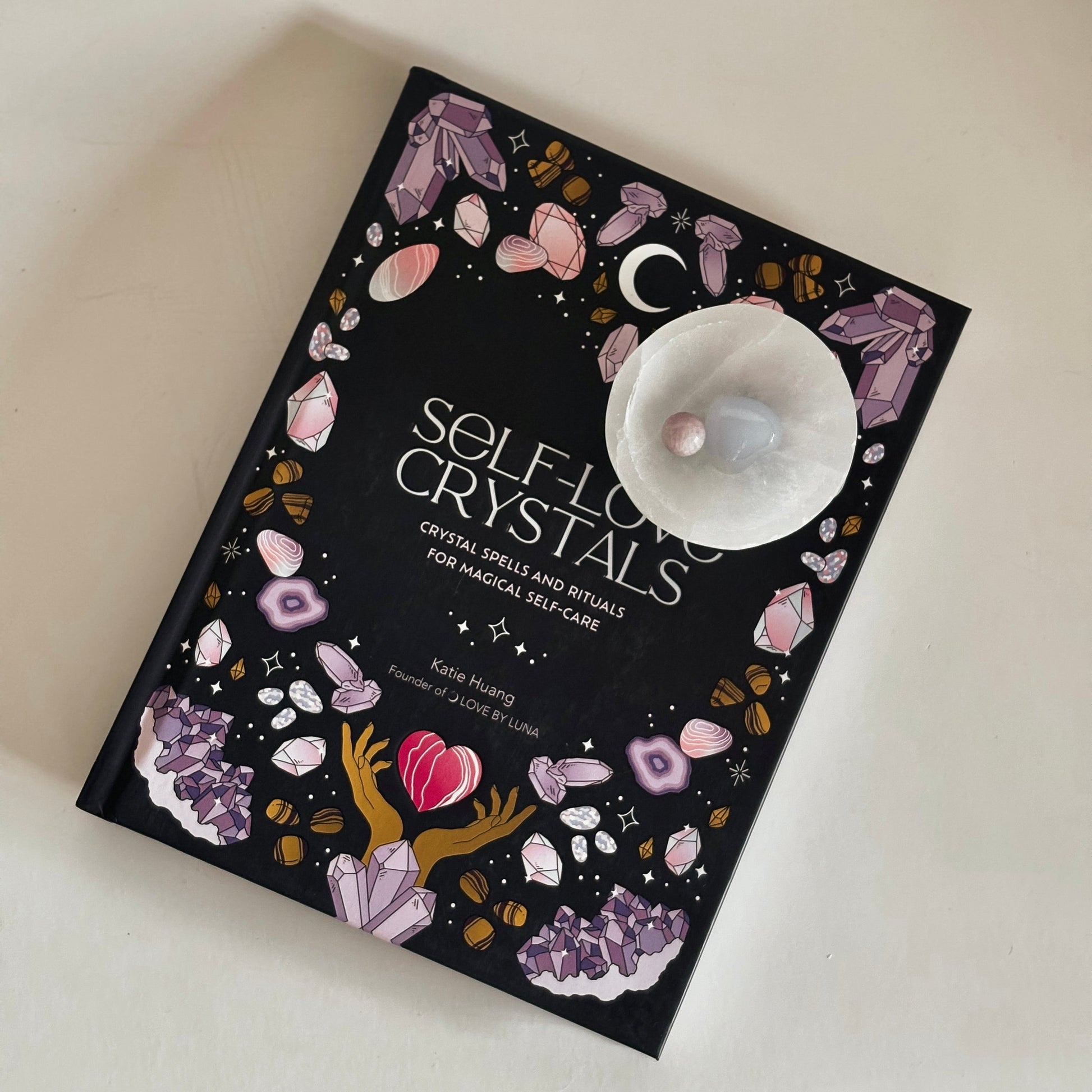 Book titled 'Self-Love Crystals' with decorative cover and selenite dish on a light background