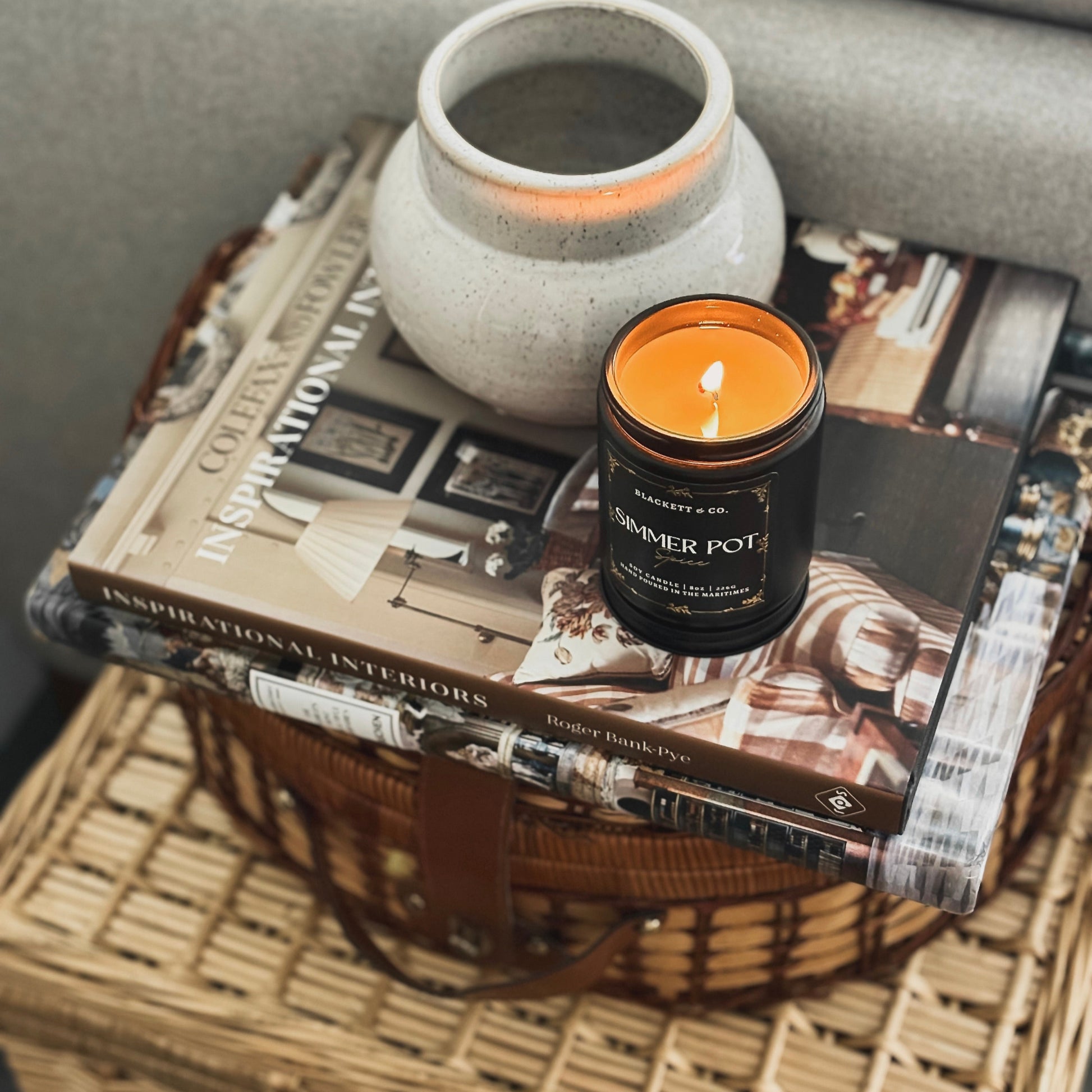 Simmer Pot Spice scented natural soy candle by Blackett & Co.burning on a stack of books and wicker baskets and ceramic container in the background.