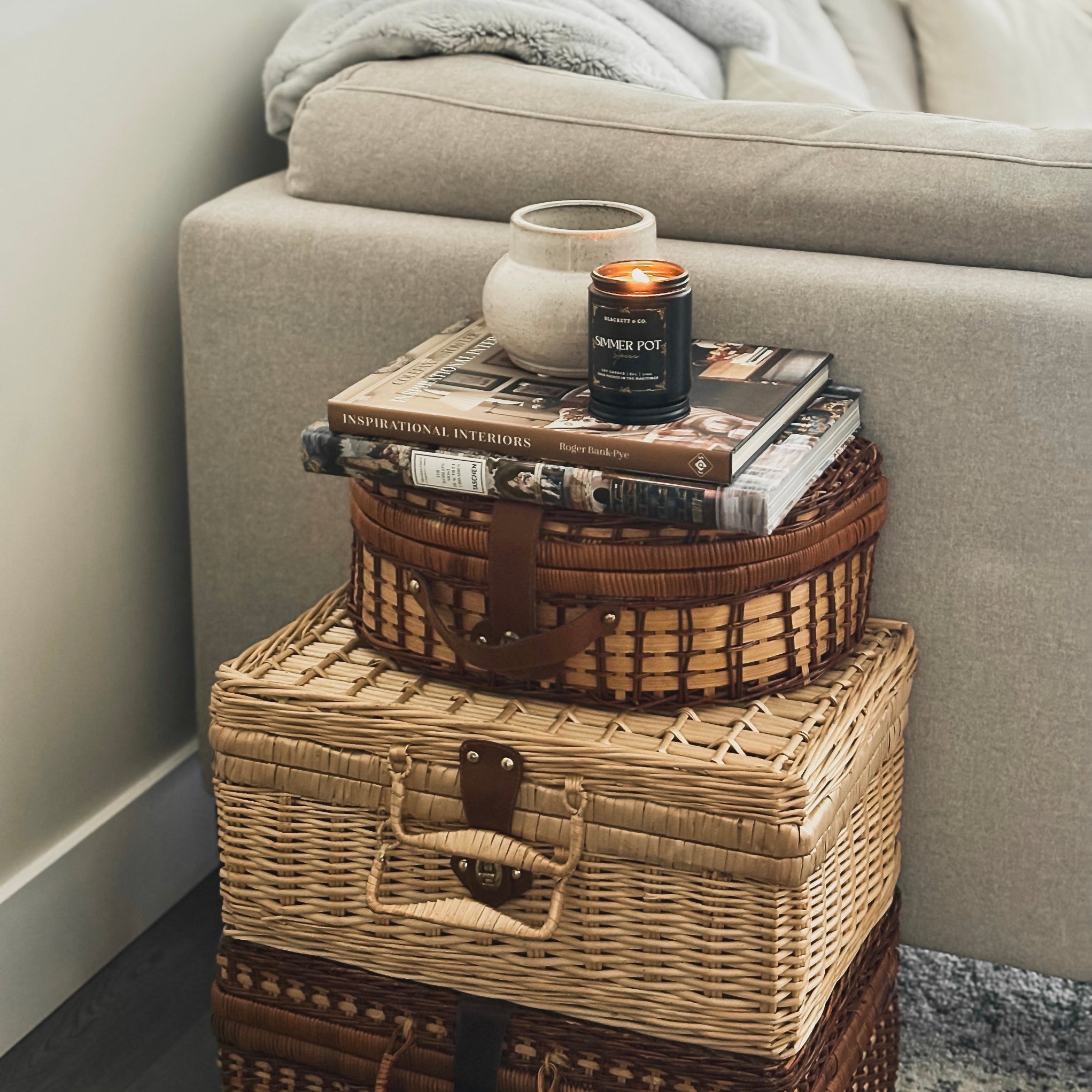 Stack of wicker baskets with a Simmer Pot Spice scented natural soy candle by Blackett & Co. and books on a sofa side table.