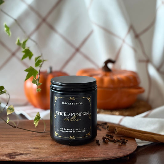 Spiced Pumpkin Hollow scented natural soy candle by Blackett & Co. on a wooden surface with orange ceramic pumpkins in the blurred background.
