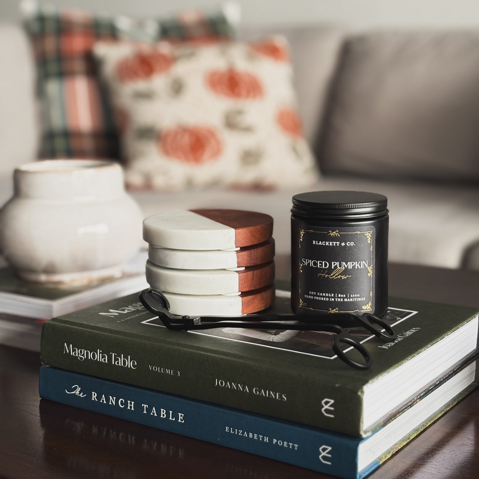 A Spiced Pumpkin Hollow Candle, books, and coasters on a coffee table with a blurred sofa  and plaid and pumpkin pillows in the background.
