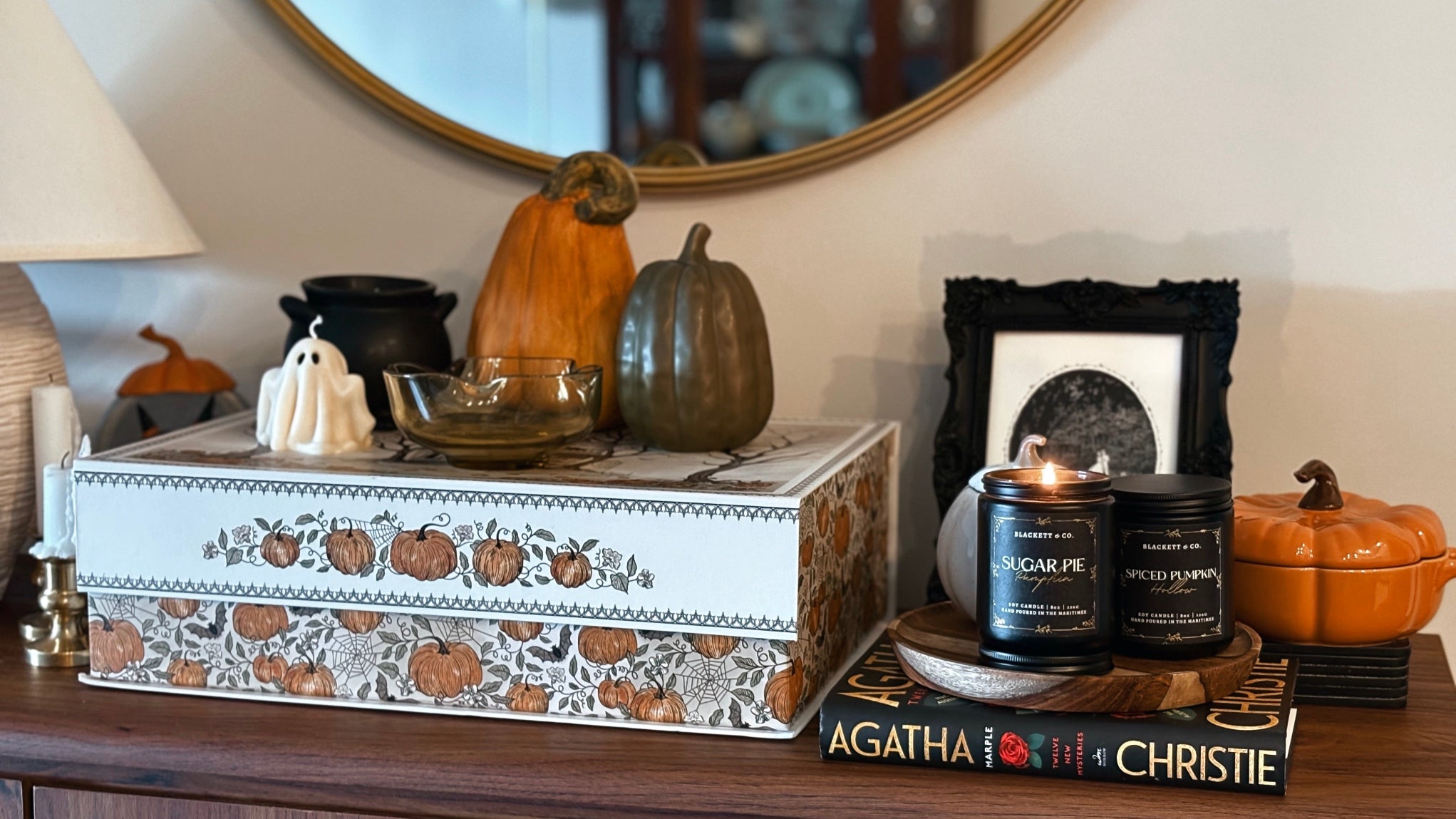 Decorative setup with pumpkins, Sugar Pie Pumpkin and Spiced Pumpkin Hollow natural scented soy candles by Blackett & Co., and a book on a table.