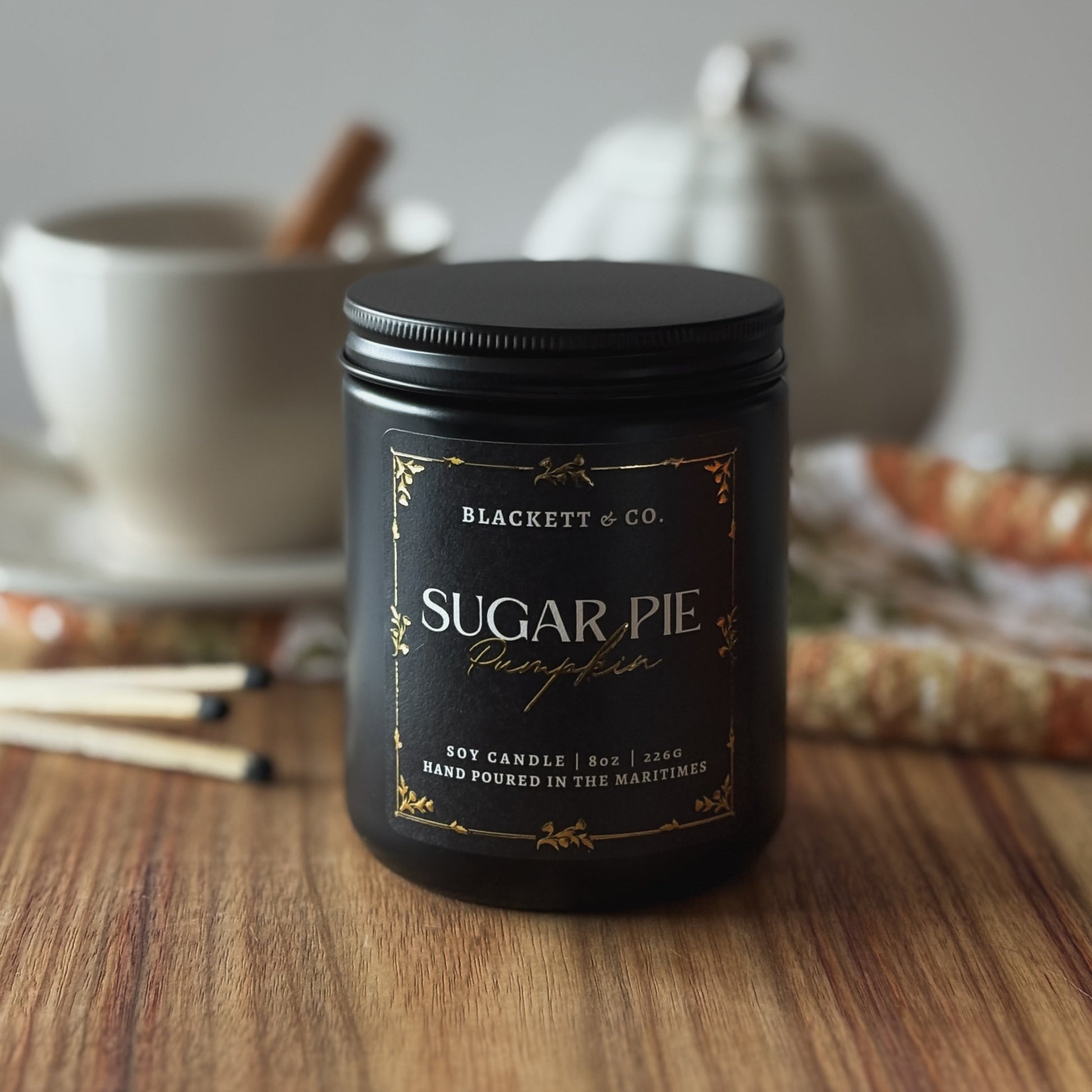 Sugar Pie Pumpkin scented natural soy candle by Blackett & Co. on a wooden surface with a white coffee cup and white ceramic pumpkin blurred background.
