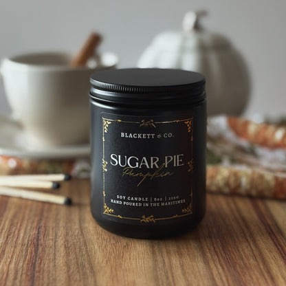 Sugar Pie Pumpkin scented natural soy candle by Blackett & Co. on a wooden surface with a white coffee cup and white ceramic pumpkin blurred background.