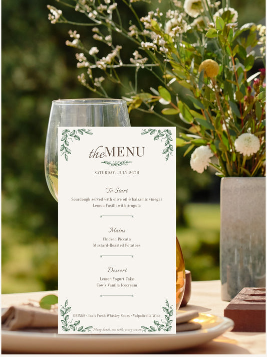 A vertical wedding menu card with green botanical illustrations and elegant typography, displayed outdoors on a table next to a wine glass and flowers.