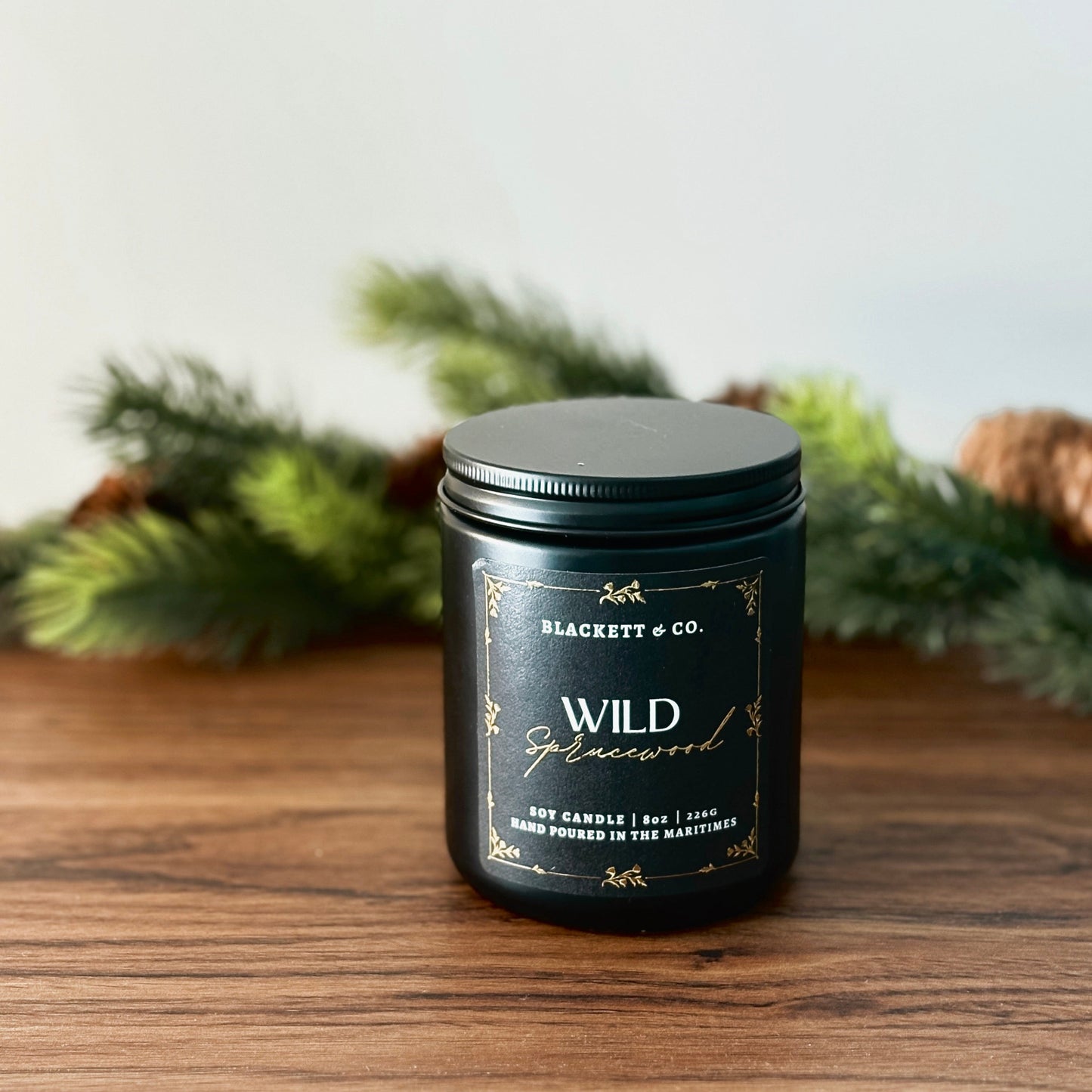 Wild Sprucewood scented natural soy candle by Blackett & Co. on a wooden surface, surrounded by greenery