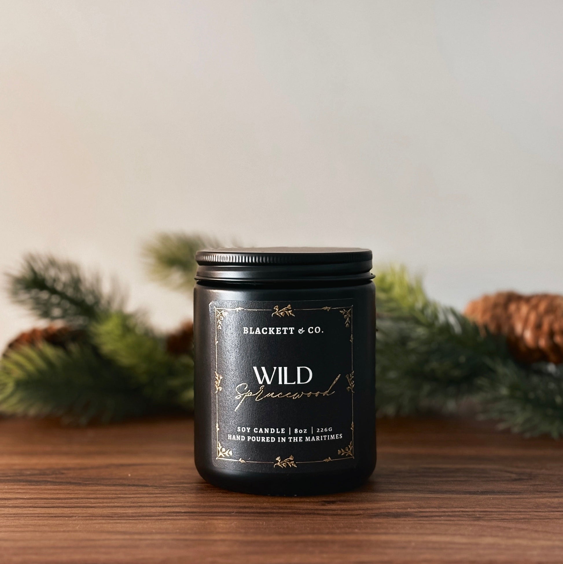 Wild Sprucewood scented natural soy candle by Blackett & Co. on a wooden surface with greenery in the background