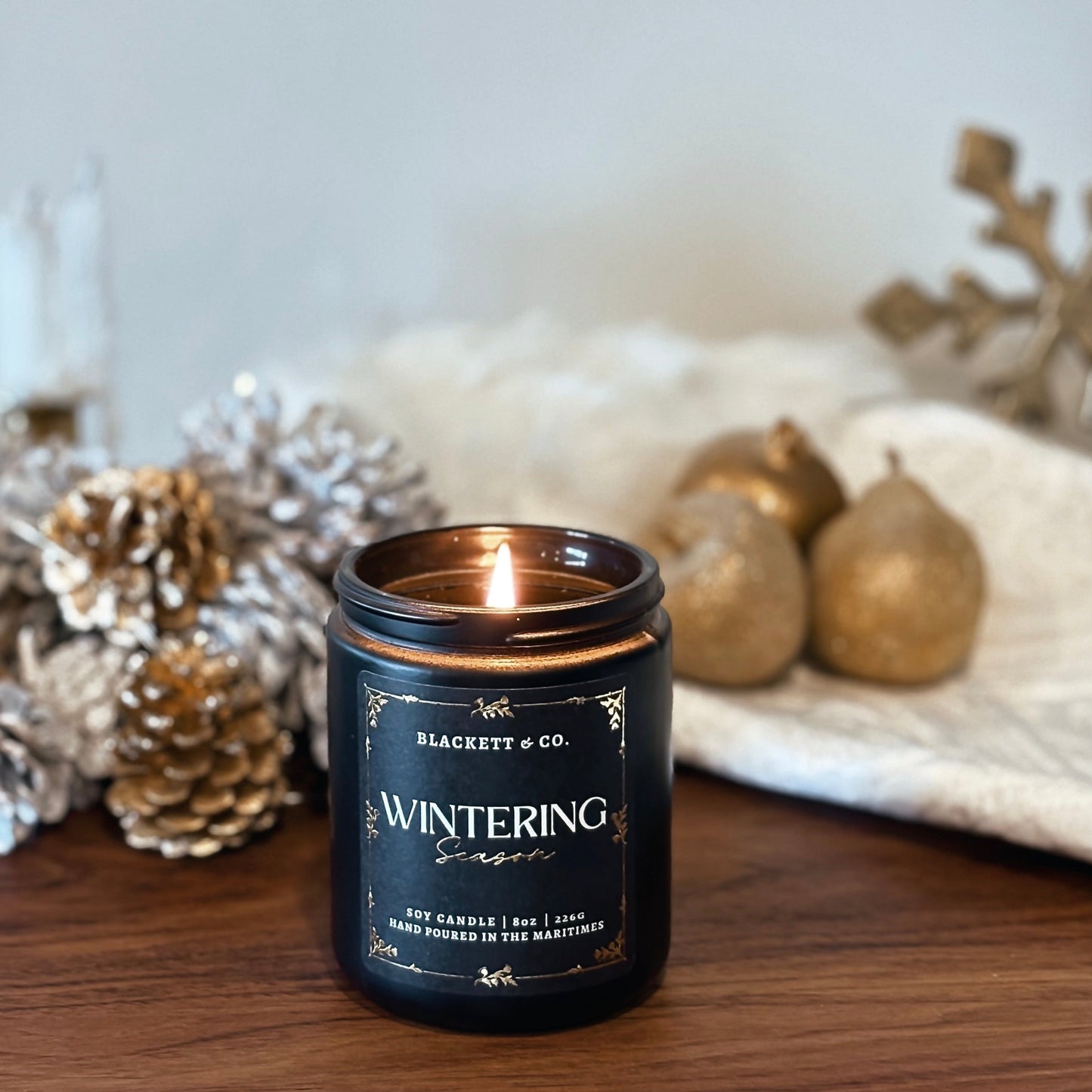 Wintering Season scented natural soy candle by Blackett & Co. on a wooden surface with frosted pine cones and decorative decor in the blurred background.