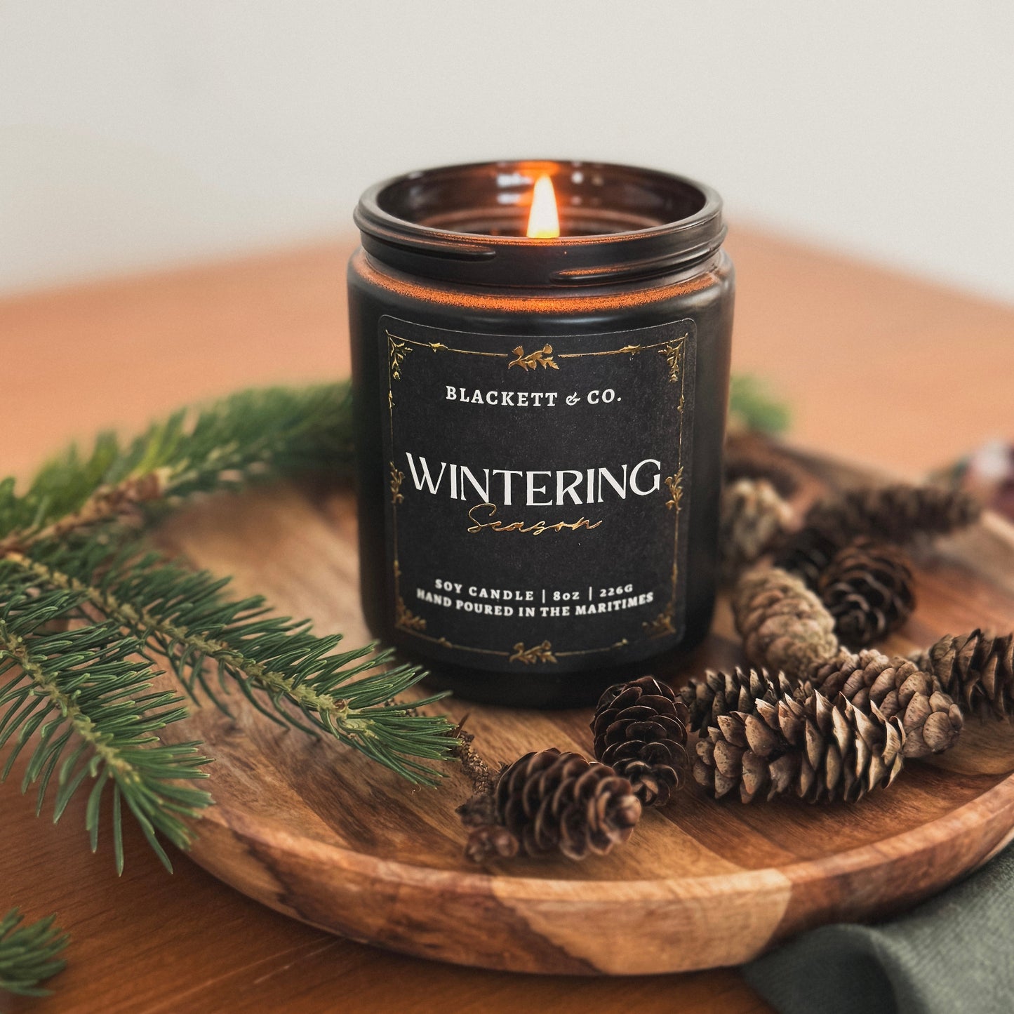 Wintering Season scented natural soy candle by Blackett & Co. on a wooden tray with pine branches and cones.